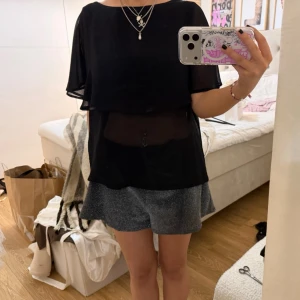 Svart transparent blus med volang - Säljer en svart blus med volangdetalj och transparent material. Blusen har korta, lösa ärmar och en avslappnad passform. Perfekt för dig som gillar en stilren och trendig look.