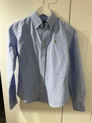 Blå skjorta från Ralph Lauren - Klassisk ljusblå skjorta från Ralph Lauren i super slim fit. Perfekt för en stilren och fräsch look. Passar tolv till trettonåringar