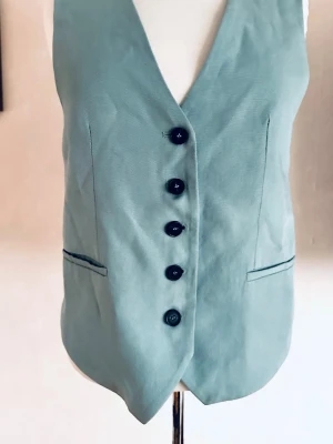 Ljusblå väst med knappar - Stilren ljusgrön väst  fem svarta knappar framtill och två fickor. Västen har en klassisk v-ringad form och är perfekt för att lyfta din outfit med en retro touch. Str Xs men är större i str