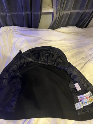 Svart pufferjacka från Moncler - Snygg svart cardigan från Moncler med stickade ärmar och detaljerad armficka med dragkedja och en moncler logga på vänster arm. Jackan har quiltad kropp och ribbade muddar. Perfekt för dig som vill ha en stilren och modern look. Den har två fickor på magen med knappar. Den har storlek 3/M 