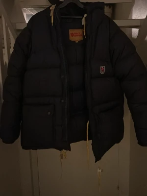 Mörkblå dunjacka från Fjällräven - Säljer en mörkblå dunjacka från Fjällräven med huva och gula dragskor. Jackan har två stora fickor framtill, klassisk Fjällräven-logga på bröstet och en stor etikett på insidan. Perfekt för kalla vinterdagar och riktigt varm.