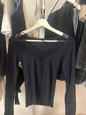 Mörkblå långärmad topp med spets - Mörkblå långärmad topp med v-ringning och dekorativ spetskant runt halsen. Toppen har en slim passform och är tillverkad i mjukt material som känns skönt mot huden. Perfekt att styla med jeans eller kjol för en trendig look.