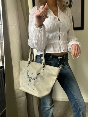 Beige handväska med örn i nitar - Snygg beige handväska i konstläder med coolt örnmotiv i silvriga nitar på framsidan. Väskan har två rejäla handtag i samma färg och metallspännen. Perfekt för dig som vill ha en unik accessoar med edgy vibe.