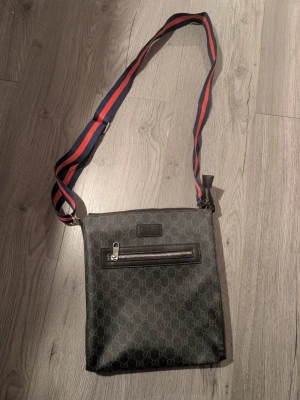 Svart axelväska - Snygg svart axelväska.. Väskan har en justerbar axelrem i blått och rött tyg som ger en sportig touch. Tillverkad i slitstarkt canvasmaterial med läderdetaljer.