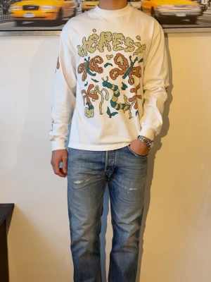 Vit långärmad t-shirt från Heresy - Vit långärmad t-shirt från Heresy med färgglad grafisk print framtill och på ärmarna. Tröjan har en relaxed passform och är tillverkad i mjuk bomull. Perfekt för dig som gillar unika prints och streetwear vibes.