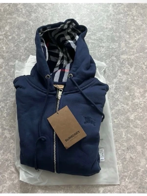 Marinblå hoodie från Burberry - Snygg marinblå hoodie från Burberry med dragkedja och klassiskt rutigt foder i huvan. Hoodien har två stora fickor framtill, ribbade muddar och justerbar dragsko i huvan. Perfekt för en stilren och avslappnad look. 