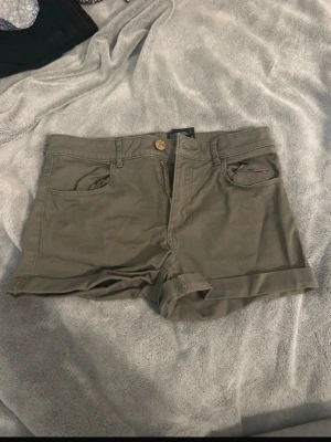 Olivgröna shorts med uppvik - Snygga olivgröna shorts med uppvikta benslut och klassisk femficksdesign. De har gylf med dragkedja och knapp, samt hällor för bälte. Perfekta för varma dagar och enkla att matcha med olika stilar.