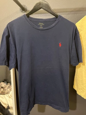 Marinblå t-shirt från Polo Ralph Lauren - Säljer en marinblå t-shirt från Polo Ralph Lauren med klassisk röd broderad logga på bröstet. T-shirten har rund hals och korta ärmar, perfekt för en avslappnad och stilren look. Materialet är mjuk bomull som känns skönt mot huden.