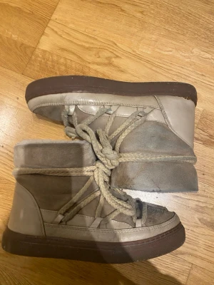 Beige moon boots från INUIKII - Säljer ett par beige moon boots från INUIKII med grova snören och mjuk, fluffig insida. Skorna har en högre modell och är tillverkade i mocka och läder med en brun sula. Perfekta för kalla dagar när du vill hålla stilen.