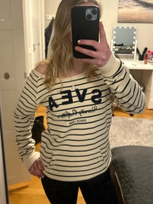 Randig tröja från Svea - Säljer en beige/vit tröja med mörkblå horisontella ränder från Svea. Tröjan har bred halsringning, långa ärmar och texten 'SVEA' i mörkblått framtill. Mjuk och skön med ribbade muddar vid ärmslut och nederkant. Perfekt för en avslappnad stil.