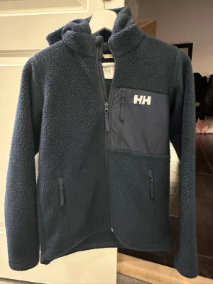 Mörkblå teddyjacka från Helly Hansen - Jag säljer en mörkblå teddyjacka från Helly Hansen i storlek 152 med huva och hel dragkedja. Jackan har två sidofickor med dragkedja och en bröstficka med HH-logga💕