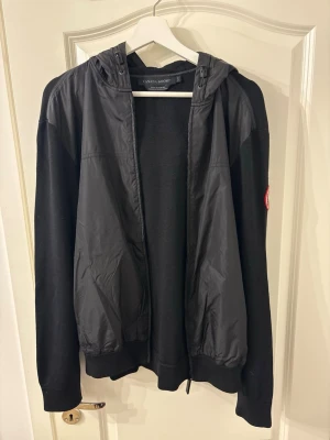 Svart hoodie från Canada Goose - Snygg svart hoodie från Canada Goose med dragkedja och huva. Jackan har ett klassiskt rött logomärke på ärmen och är tillverkad i en mix av tyg och glansigt material framtill. Perfekt för dig som vill ha en stilren och bekväm look.