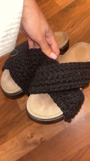 Svarta stickade sandaler med korsad rem - Säljer ett par sandaler med breda, korsade remmar i svart stickat material. Sulan är beige och ser riktigt bekväm ut, perfekt för sommardagar. Sandalerna har en avslappnad och trendig look som passar till många outfits.