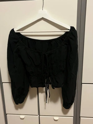 Svart blus med snörning - Trendig svart blus med långa puffärmar och snörning framtill. Blusen har en croppad passform och är gjord i ett lätt tyg som ger en avslappnad men snygg look. Perfekt att styla med höga jeans eller kjol.