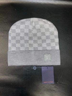 Grå Louis Vuitton mössa med rutmönster - Snygg grå mössa från Louis Vuitton med klassiskt rutmönster och vikt kant. LV-logga på framsidan och diskret etikett. Tillverkad i mjukt stickat material som känns lyxigt och stilrent. Perfekt accessoar för dig som gillar exklusiv streetwear.