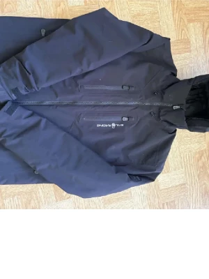 Svart vindjacka Sail Racing - Svart vindjacka från Sail Racing med huva och dragkedja framtill. Jackan har två bröstfickor med dragkedja och logga på bröstet. Stilren design och praktiska detaljer, perfekt för blåsiga dagar. Finns lite små hål 