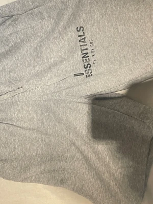 Grå mjukisbyxor Essentials Fear of God - Säljer ett par grå mjukisbyxor från Essentials Fear of God. Byxorna har en relaxed fit, dragsko i midjan och tryckt logga på benet. Tillverkade i mjukt bomullsmaterial som är perfekt för chill dagar. Snygg streetwear-vibe och riktigt bekväma.