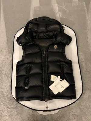 Svart moncler bormes vest - Hör av innan köp! Bästa på marknaden. Skön inför sommaren, riktig brud magnet. Snygg svart dunväst från Moncler med avtagbar huva och två dragkedjefickor framtill. Västen har klassisk Moncler-logga på bröstet och quiltad design. Perfekt för lager-på-lager och håller dig varm med stil. Fodrad insida och coola detaljer.