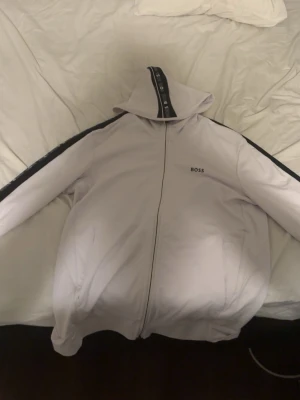 Vit hoodie från BOSS - Snygg ljuslila hoodie från BOSS med dragkedja framtill och svarta detaljer längs ärmarna. Märket syns tydligt på bröstet och huvan har en cool logotypremsa. Perfekt för en avslappnad och trendig look.