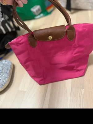 Rosa handväska från Longchamp - Snygg rosa handväska från Longchamp med bruna handtag och lock i äkta läder. Väskan har klassisk design med guldfärgad knapp och är tillverkad i slitstark nylon. Perfekt för dig som vill ha en färgklick till din outfit.