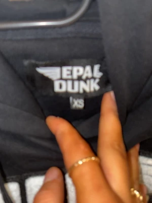 Svart EPA DUNK hoodie med tryck - Säljer en svart hoodie från EPA DUNK i storlek XS. Hoodien har ett stort vitt tryck med EPA DUNK-loggan på bröstet, klassisk huva med dragsko och en stor magficka framtill. Perfekt för dig som gillar streetwear och vill ha en skön och stilren look.
