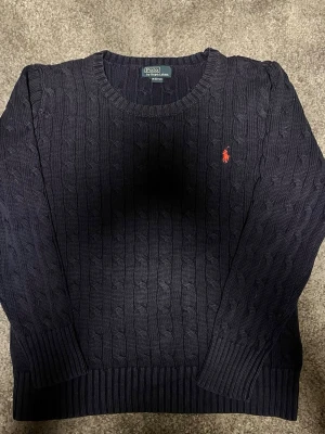 Polo Ralph Lauren - Mörkblå kabelstickad tröja från Polo Ralph Lauren med rund halsringning och röd broderad logga på bröstet. Tröjan har långa ärmar och ribbade muddar vid ärmslut och nederkant. Perfekt för en stilren och klassisk look.