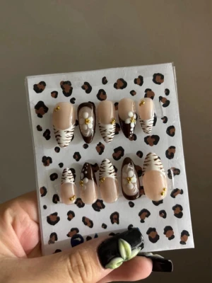 Bruna press-on naglar med zebramönster - Snygga press-on naglar i mandelform med beige bas, bruna och vita zebramönster, 3D-blommor och små guldfärgade detaljer. Perfekt för dig som vill ha en trendig och unik look. Tillverkade i plast. 10st press-ons + gåva!