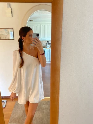 Studentklänning - Säljer en vit kortklänning med oneshoulder-design och en lång puffärm. Polyester men sidenkänsla. Storlek 36 från NAKD, använd endast en kort stund under studenten, annars aldrig använd. 