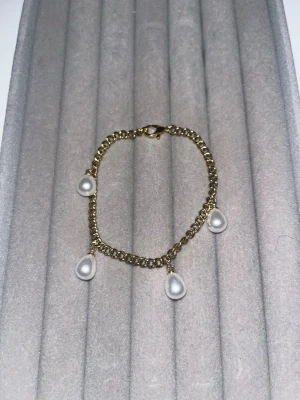 Guldfärgat armband med droppformade pärlor - Snyggt armband i guldfärgad metall med fyra droppformade vita pärlor som hänger längs kedjan. Perfekt accessoar för att ge din outfit en elegant touch. Lätt att matcha med andra smycken och har ett klassiskt karbinlås.