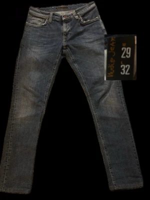 Blå  jeans från Nudie Jeans - Säljer ett par klassiska blå jeans från Nudie Jeans med femficksmodell. Jeansen har snygga slitningar och kontrastsömmar, samt Nudie-loggan på bakfickan. Tillverkade i slitstark denim som ger en tidlös look.