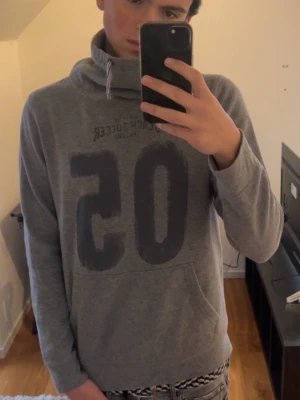 Grå hoodie med tryck och snörning från Spirit  - Säljer en grå hoodie med stort tryck '05' på framsidan och texten 'Beach Soccer Challenge'. Tröjan har en hög krage med snörning och en stor magficka. Det är en neckwarmer, och har inte en hood. Storleken är en M. Modellen i bilden är 188cm lång och väger 74kg. 