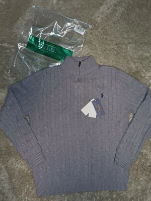 Grå kabelstickad tröja Polo Ralph Lauren - Snygg grå kabelstickad tröja från Polo Ralph Lauren med halv dragkedja och hög krage. Tröjan har det klassiska blå logobroderiet på bröstet och ribbade muddar vid ärmslut och nederkant. Perfekt för lager på lager och stilren vardagslook.