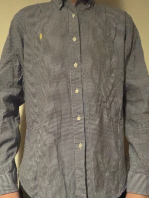 Blårutig skjorta från Polo Ralph Lauren - Klassisk blårutig skjorta från Polo Ralph Lauren med vit knappstängning och gul broderad logga på bröstet. Skjortan har lång ärm och är tillverkad i bomull. Perfekt för en stilren look. Storlek M-L men mer M