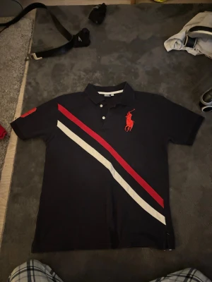 Svart pikétröja från Ralph Lauren - Svart pikétröja från Ralph Lauren med röd logga på bröstet och diagonala ränder i rött och vitt framtill. Klassisk krage, knappar vid halsen och korta ärmar. Perfekt för en stilren och sportig look.