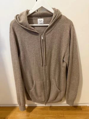 Beige cashmere hoodie John Henric - Mjuk och lyxig beige hoodie från John Henric i 100% cashmere. Dragkedja framtill, två fickor och ribbade muddar. Klassisk huva med dragsko för extra komfort. Perfekt för dig som vill ha en stilren och bekväm hoodie i premiumkvalitet.