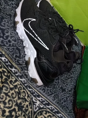 Nike React vision  - Snygga svarta Nike react sneakers med vit, vågig sula och stora vita swoosh-loggor på sidorna. Ovandelen är i mesh-liknande material för bra ventilation och skorna har svarta skosnören. Sulan har detaljer i svart och brunt för en cool kontrast.