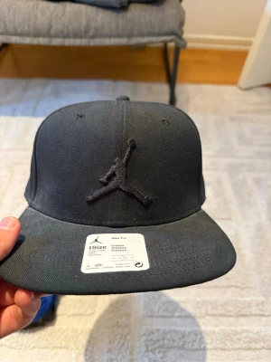 Svart Jordan keps med broderad logga - Snygg svart keps från Jordan med den klassiska broderade Jumpman-loggan framtill. Kepsen har rak skärm och justerbar baksida. Tillverkad i bomullsmix och har texten 'Quality inspired by the greatest player ever' på insidan. Perfekt för en sportig och streetwear-inspirerad stil.
