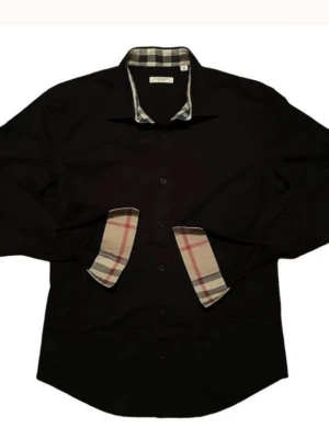 Svart skjorta med rutiga detaljer - Stilren svart skjorta från Burberry med klassiska rutiga detaljer på insidan av kragen och manschetterna. Skjortan har lång ärm, knappar framtill och en tidlös design som passar till många tillfällen.   Skicket är näst intil helt ny då den endast har testats och används en enstaka dag samt att den har tvättats endast en gång på kemtvätt    Såklart äkta köpt från en Burberry butik Fler bilder kan skickas vid seriöst intresse  (kan mötas upp i Göteborgs trakterna)