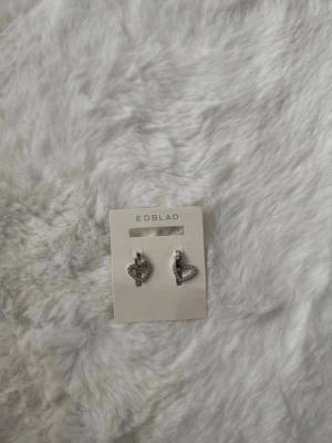 Hjärta örhängen från Edblad - Snygga örhängen i form av hjärtan från Edblad. De är silvriga och dekorerade med små gnistrande stenar som ger en lyxig känsla. Tillverkade i rostfritt stål och perfekta för att lyfta din stil med en söt detalj.