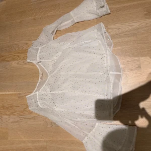 Vit transparent blus med guldprickar - Säljer en superfin vit blus från Hollister i transparent material med små guldprickar över hela tyget. Blusen har lång ärm och en lös, flowy passform som ger en luftig känsla. Perfekt att styla med jeans eller kjol för en trendig look.