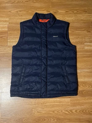 Mörkblå dunväst från GANT - Snygg mörkblå dunväst från GANT med quiltad design och ståkrage. Västen har två dragkedjeförsedda fickor på framsidan och diskret logga på bröstet. Perfekt för lager-på-lager och är perfekt för en liten kallare sommar dag