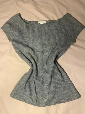 Grå ribbad topp från H&M - Säljer en grå ribbad topp från H&M i storlek M. Toppen har en bred halsringning och är kortärmad med en mjuk, stretchig passform. Perfekt att styla med jeans eller kjol för en enkel och snygg look. 