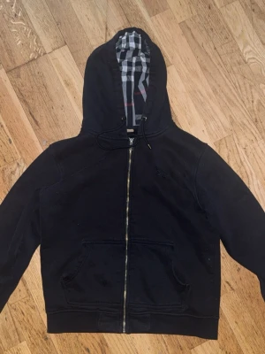 Svart hoodie med dragkedja Burberry - Säljer en svart hoodie från Burberry med dragkedja och klassiskt rutigt foder i huvan. Tröjan har två stora fickor framtill och diskret logga på bröstet. Perfekt för dig som gillar stilrena och exklusiva plagg. Stroleken är L men jag skulle nog klassa den som en M passar perfekt för M och även S! 
