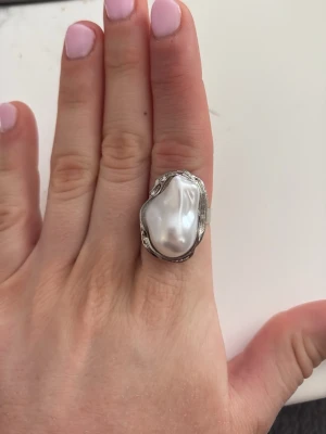 Statementring med stor pärla - Unik silverfärgad ring med en stor, oregelbunden pärla som centerpiece. Ringen har en bred, detaljerad ringbas i metall som ger ett coolt och trendigt intryck. Perfekt för dig som vill sticka ut med ett smycke som verkligen syns. Har köpt från en secondhand butik, runt storlek 17/18 men går att justera. Använd ett fåtal gånger. Tror inte de är äkta silver, men vet inte om den rostar
