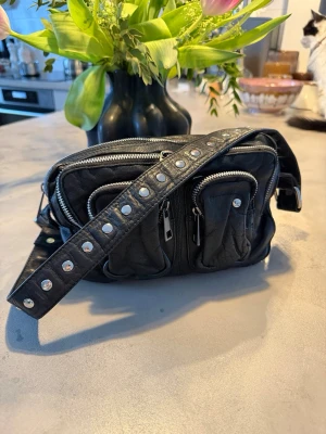 Svart axelväska med nitar och dragkedjor NUNOO - Cool svart axelväska i skinnimitation med bred axelrem dekorerad med silverfärgade nitar från NUNOO. Väskan har flera fack med dragkedjor framtill och på toppen, perfekt för att hålla ordning på småsaker. Snygg och edgy design som sticker ut.