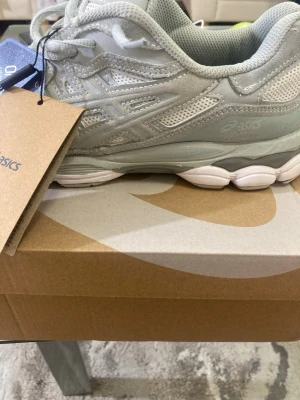 Ljusgrå sneakers från Asics - Säljer ett par stilrena ljusgrå Asics sneakers med mesh och mockadetaljer. Skorna har snörning, vit och grå sula samt klassisk Asics-logga på sidan. Perfekta för dig som gillar sportig och clean stil.