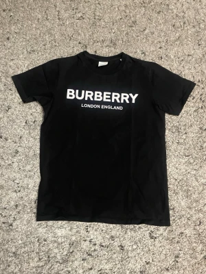 Svart Burberry t-shirt med logga - Svart t-shirt från Burberry med vit logotyp och texten 'London England' på bröstet. Klassisk rund hals och korta ärmar. Tillverkad i mjuk bomull som känns skön mot huden. Perfekt för en clean och stilren look.