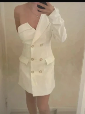 Vit oneshoulder kavajklänning - Stilren vit kavajklänning med oneshoulder-design och dubbelknäppning framtill. Klänningen har dekorativa fickor och en asymmetrisk axel, vilket ger en trendig och unik look. Perfekt för dig som vill sticka ut med en modern twist.