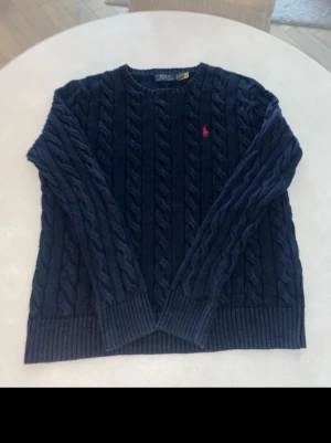 Mörkblå kabelstickad tröja Polo - Mörkblå kabelstickad tröja från Polo Ralph Lauren med klassisk rund hals och ribbade muddar. Tröjan har ett rött broderat Polo-logo på bröstet och är gjord i mjukt material som passar perfekt för kyliga dagar.