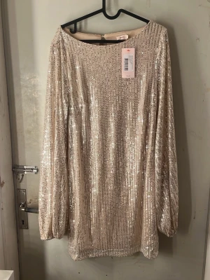 Champagnefärgad paljettklänning - Säljer en glittrig champagnefärgad kortklänning med paljetter och långa ärmar. Klänningen har en rak passform och en snygg öppning i ryggen. Perfekt för dig som vill sticka ut på festen! Helt ny oanvänd, prislapp kvar